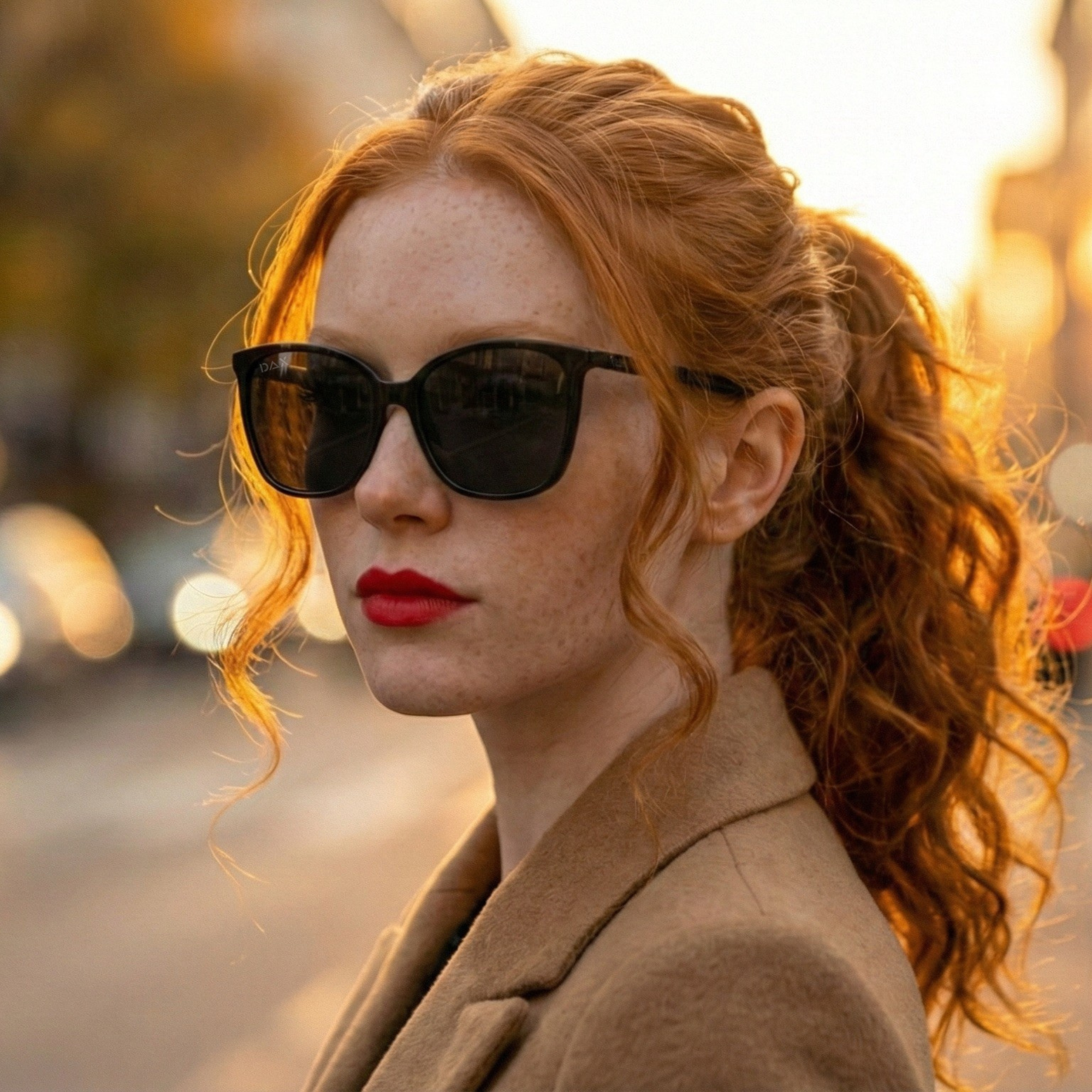 Woman with red hair wearing sunglasses and a beige coat outdoors