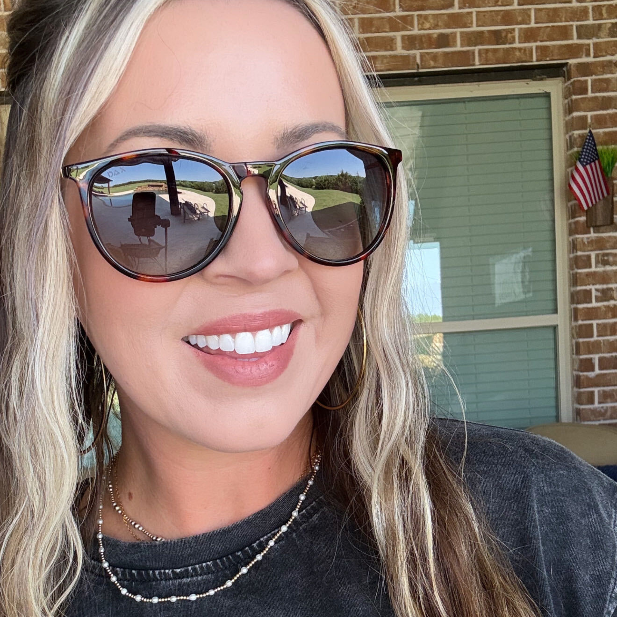A close-up selfie of a woman wearing large tortoiseshell sunglasses with dark tinted lenses, standing outdoors in front of a brick wall and window.