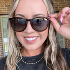A close-up lifestyle shot of a woman wearing cat-eye sunglasses with tortoiseshell frames and dark lenses, adjusting the side temple with her hand. She is wearing a black t-shirt and a gold beaded necklace, with a brick wall and window in the background.