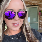 A close-up lifestyle shot of a woman wearing round sunglasses with tortoiseshell-patterned frames and vibrant purple mirrored lenses, set against a background of a brick wall and window.