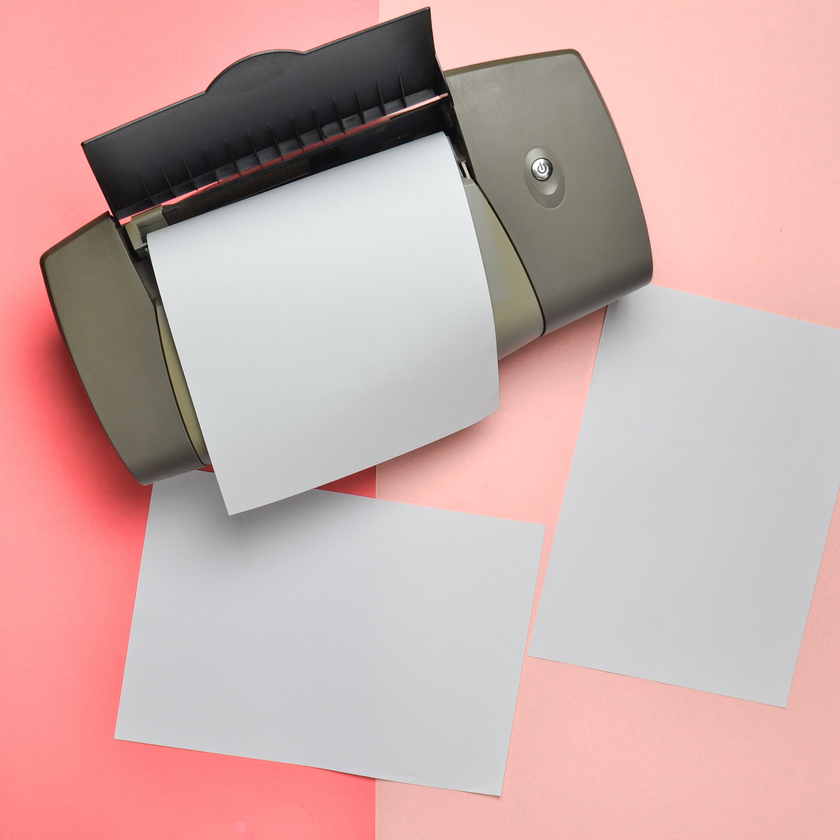 A compact gray and black paper roll printer with a white sheet emerging from the opening, set on a pink background with two blank white sheets nearby.