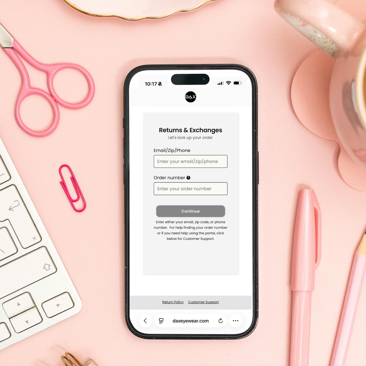 A smartphone centered on a pink desk flat lay, displaying a returns and exchanges webpage. The phone is surrounded by pink scissors, a white keyboard, a pink paperclip, a pink pen, and pastel stationery items.