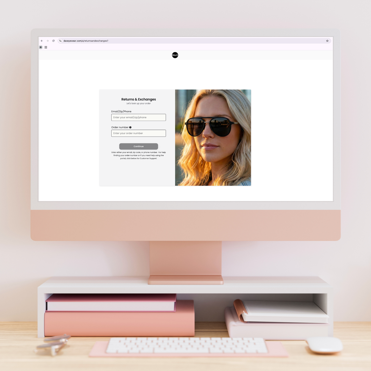 A desktop computer monitor on a stand with a keyboard, mouse, and books on a desk, all in a soft blush pink color scheme. The computer screen displays a website login page with a photo of a woman wearing sunglasses.