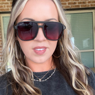 A close-up of a woman wearing black square sunglasses with dark red tinted lenses and a visible 'DAK' brand mark on the left lens, set against a background of a brick wall and window.