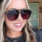 A close-up of a woman wearing black aviator-style sunglasses with dark gradient lenses and the brand name DAX visible on the left lens frame.