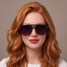 A close-up portrait of a woman with red hair and freckles wearing dark sunglasses with a black frame and dark purple lenses, set against a solid taupe background.