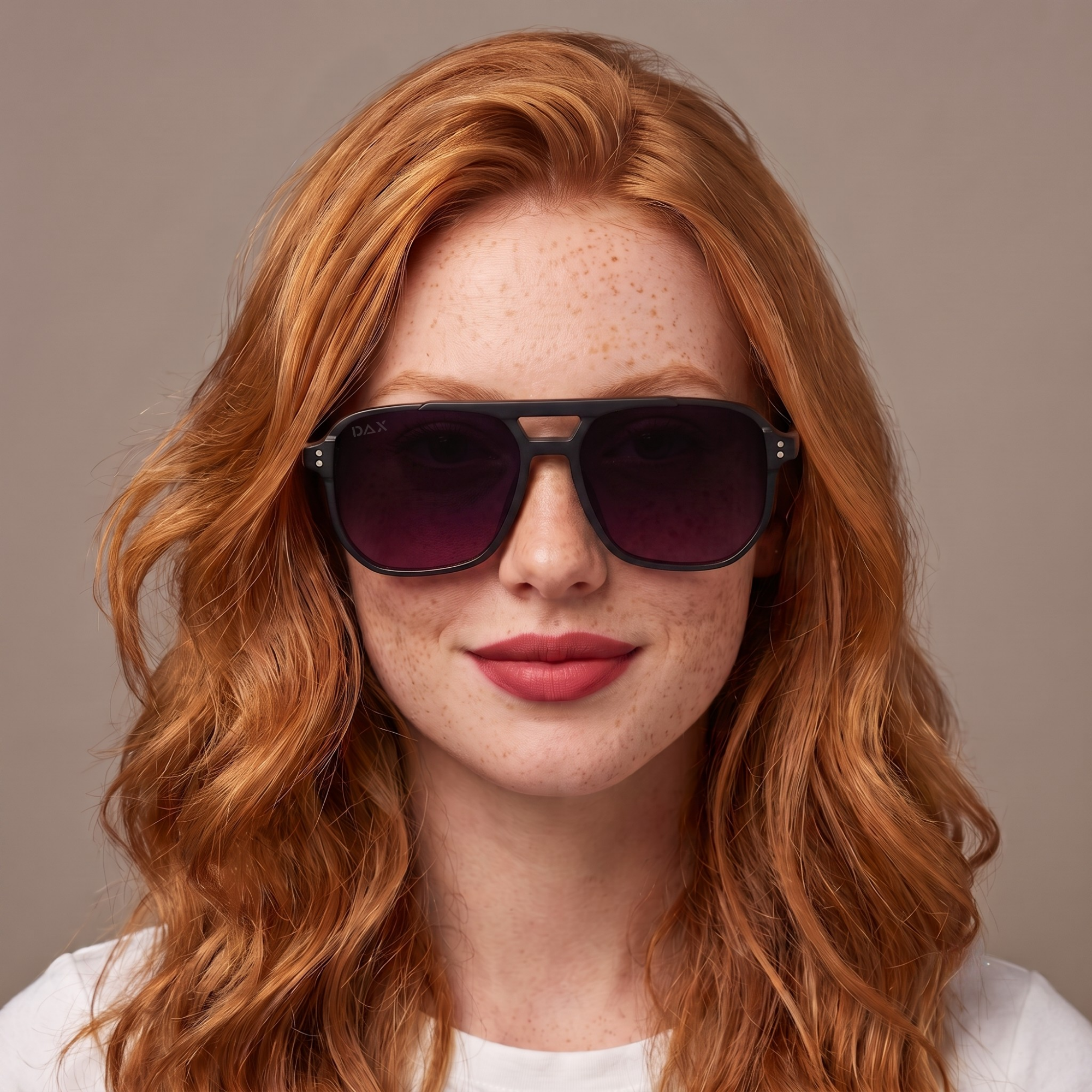 A close-up portrait of a woman with red hair and freckles wearing dark sunglasses with a black frame and dark purple lenses, set against a solid taupe background.