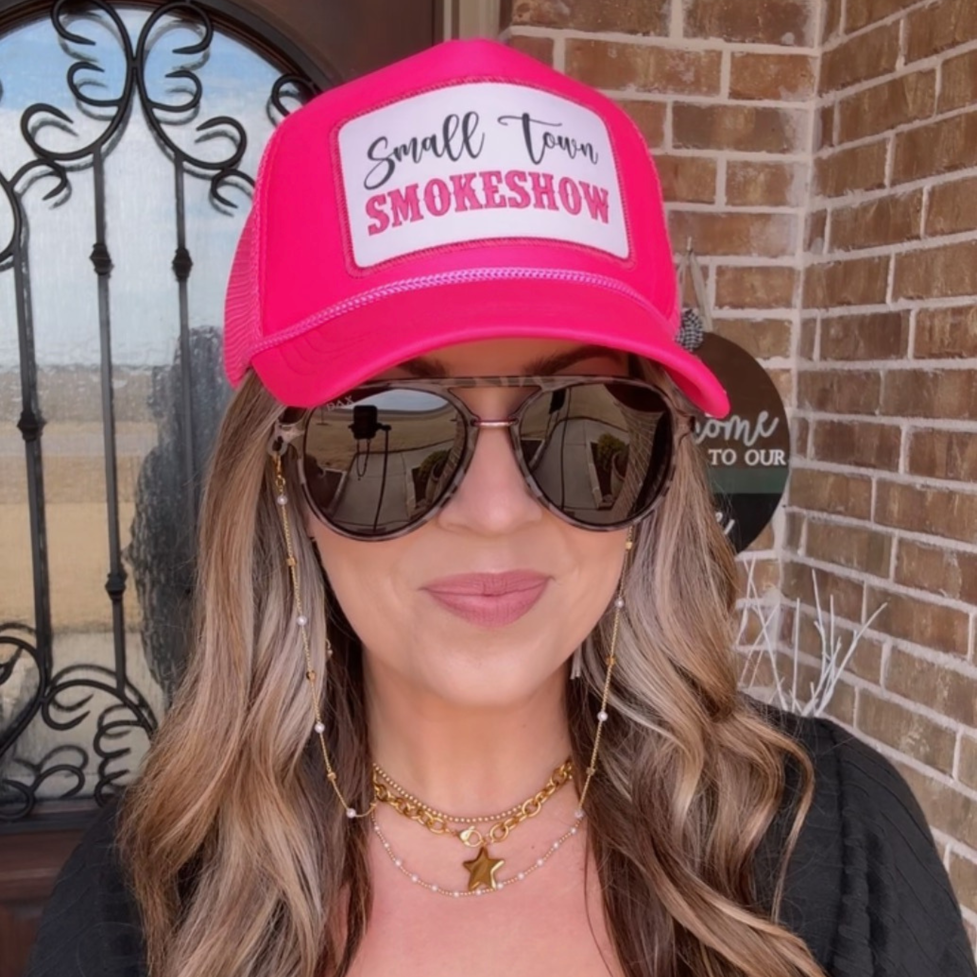 Person wearing a pink cap with 'Small Town Smokeshow' text, sunglasses, and layered necklaces.