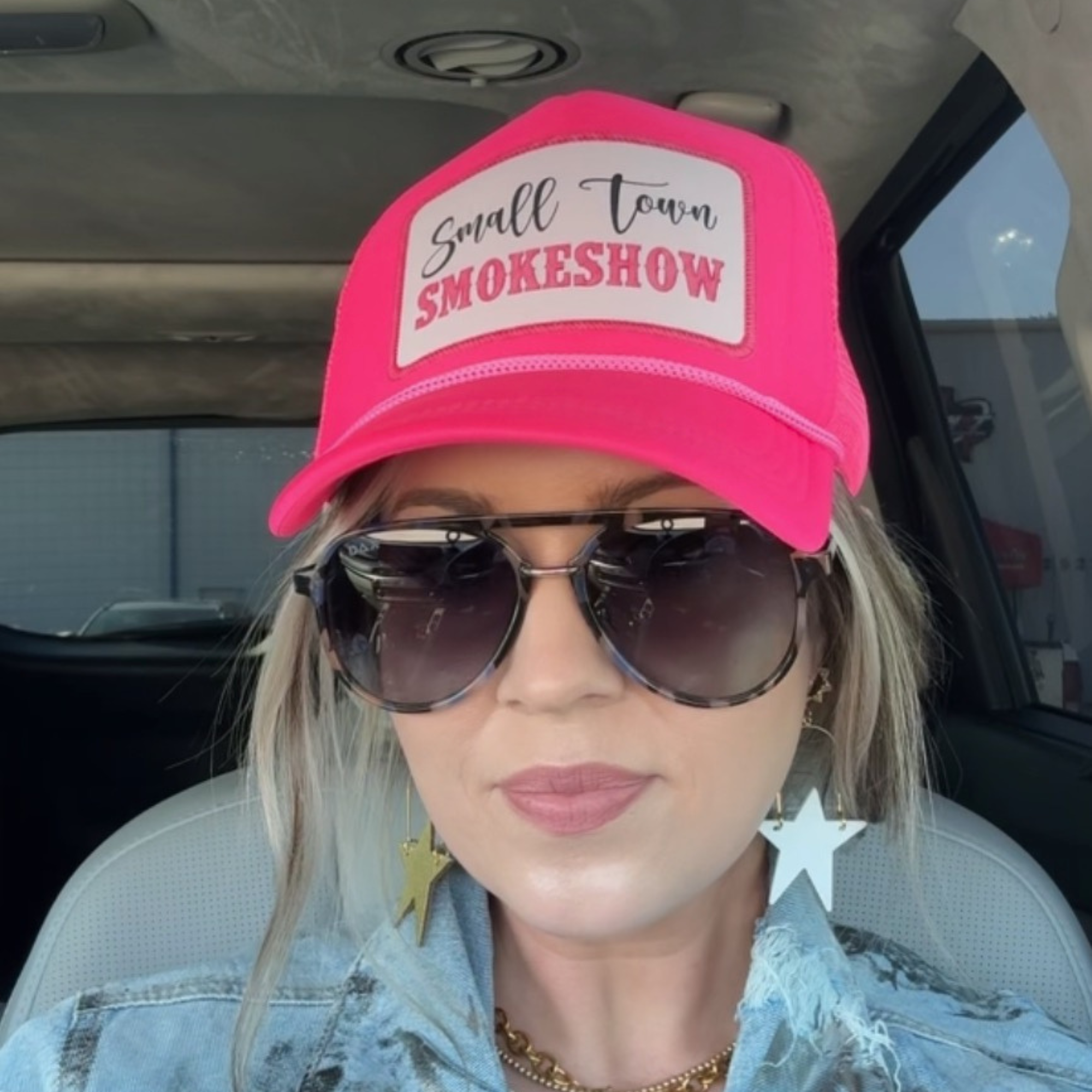 Person wearing a pink cap with 'Small Town Smokeshow' text and sunglasses inside a vehicle.