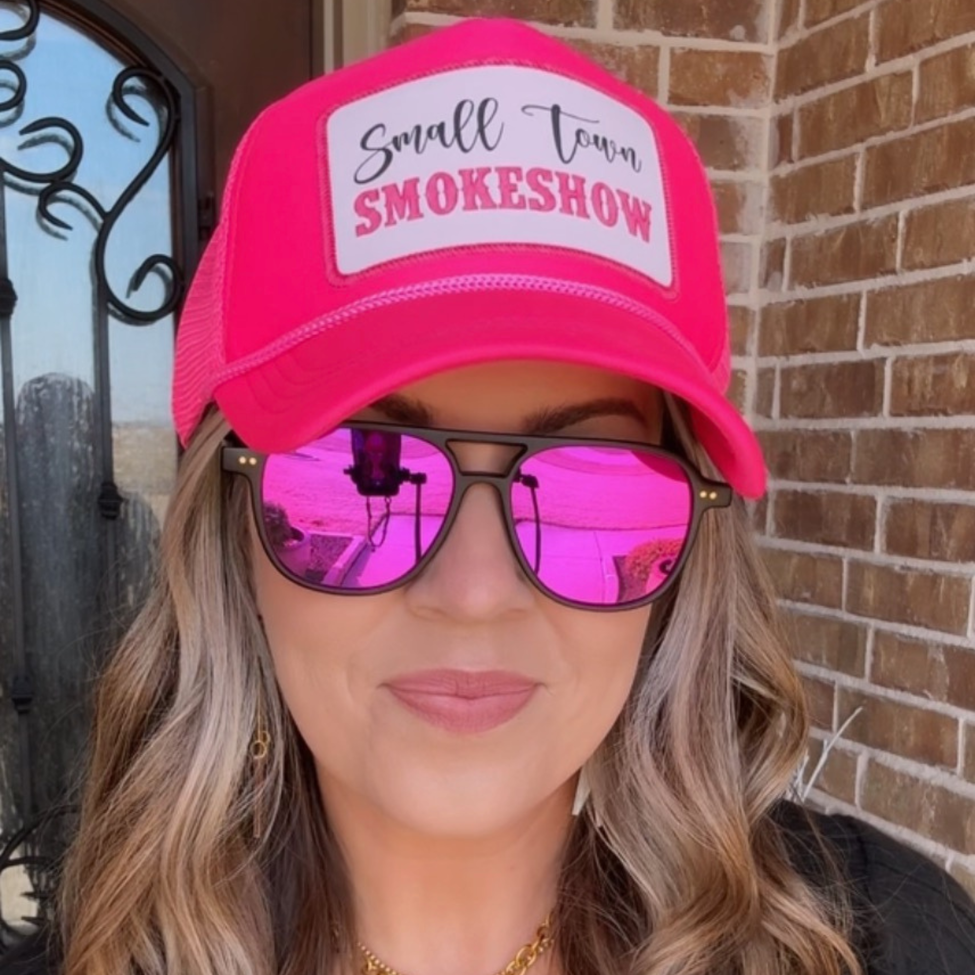 Person wearing a pink cap with 'Small Town Smokeshow' text and pink sunglasses, standing in front of a brick wall.