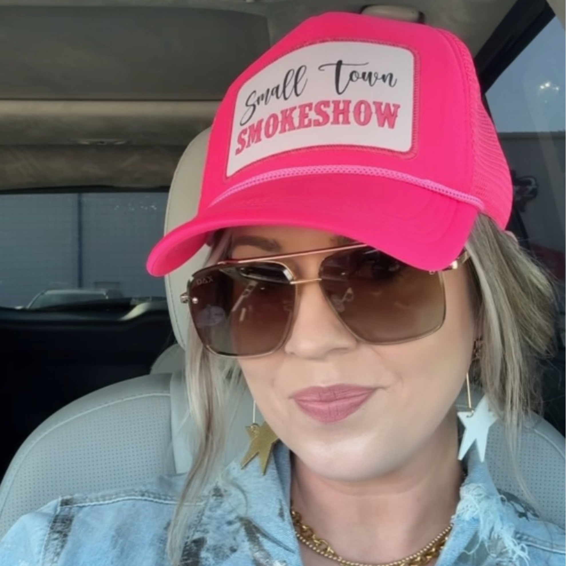Person wearing a pink cap with 'Small Town Smokeshow' text and sunglasses, sitting inside a vehicle.