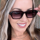 A close-up of a woman wearing dark brown gradient sunglasses with a thick black frame, holding one lens with her finger.
