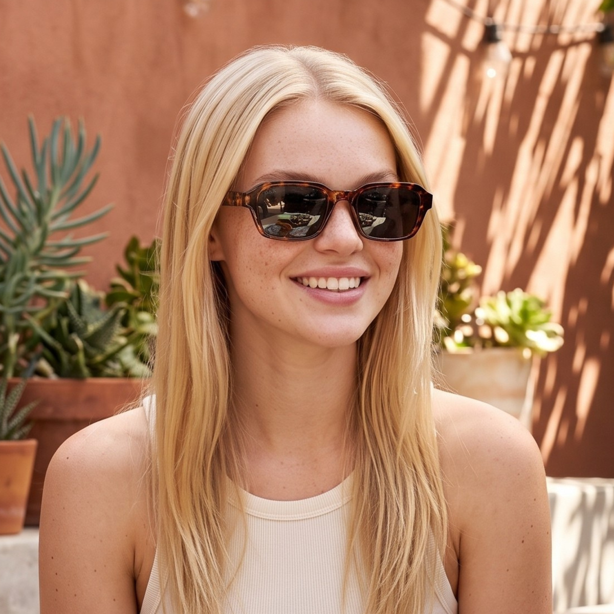 A smiling blonde woman wearing tortoiseshell square sunglasses with dark lenses, photographed outdoors against a warm terracotta wall with plants in the background.