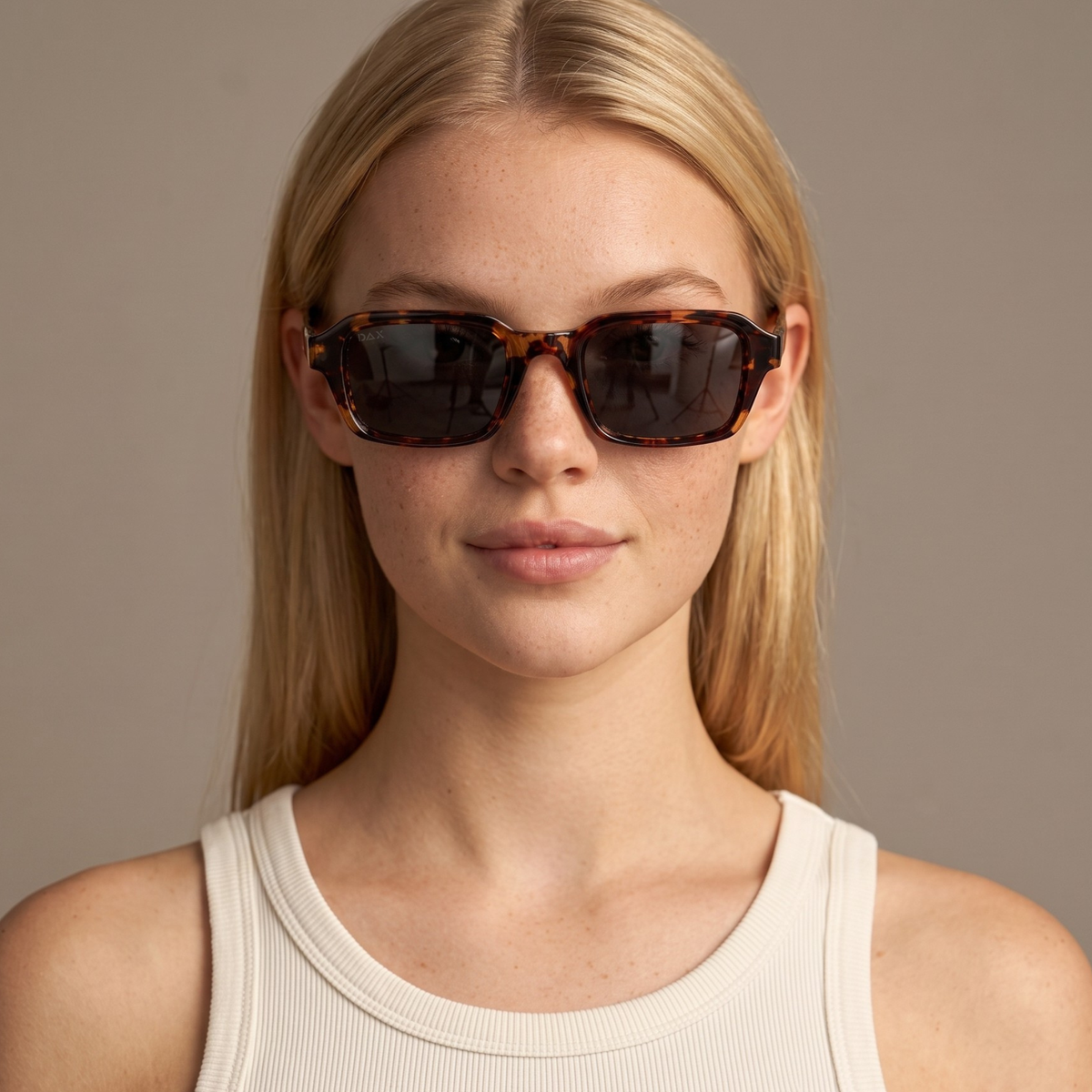 A close-up of a blonde woman wearing rectangular tortoiseshell sunglasses with dark lenses and a ribbed cream tank top, set against a solid warm gray background.